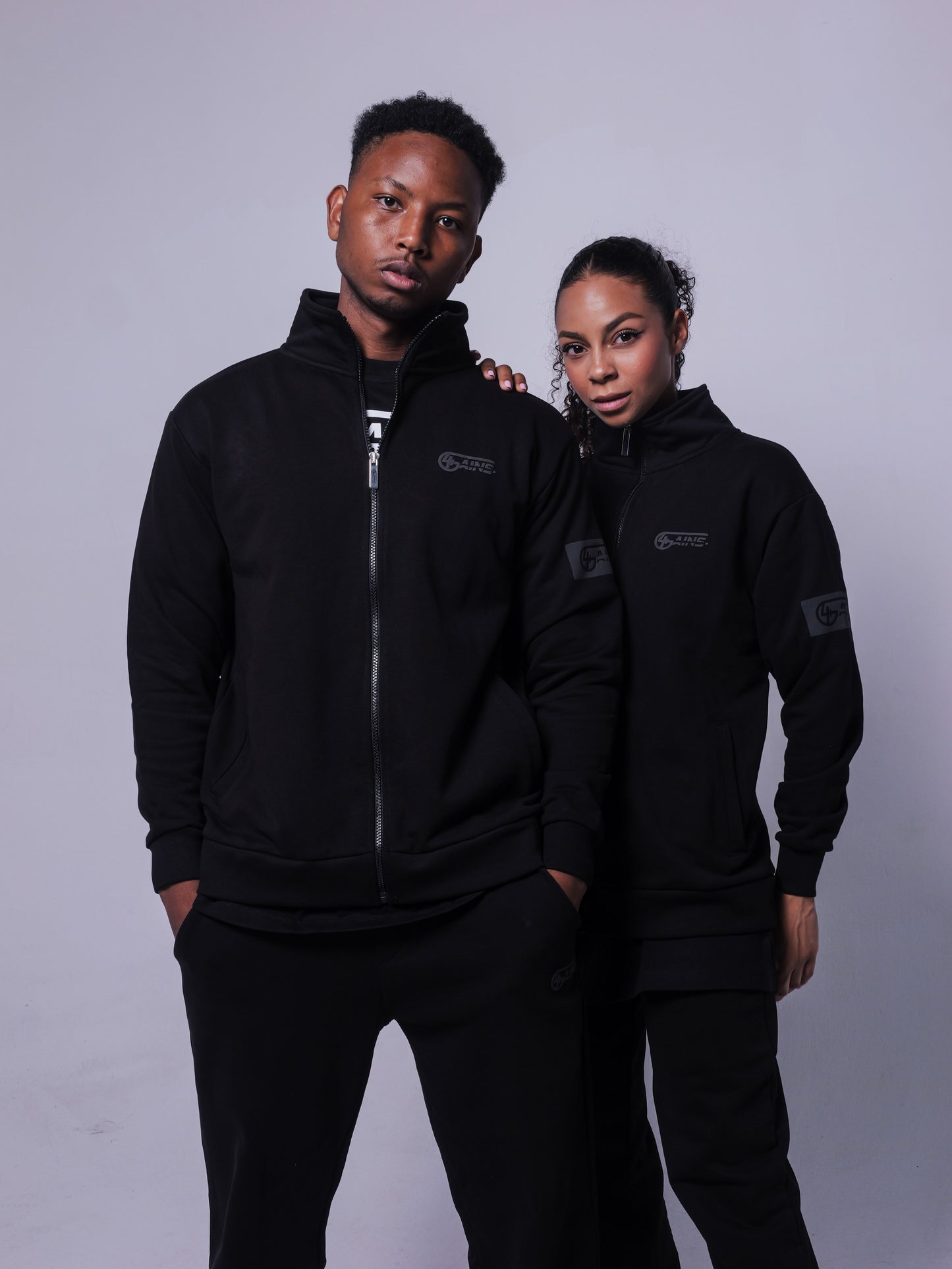 4GAINS Tracksuit Jacke in der Farbe schwarz mit schwarzem Puff Print an einem stehenden weiblichen und männlichem Model 
