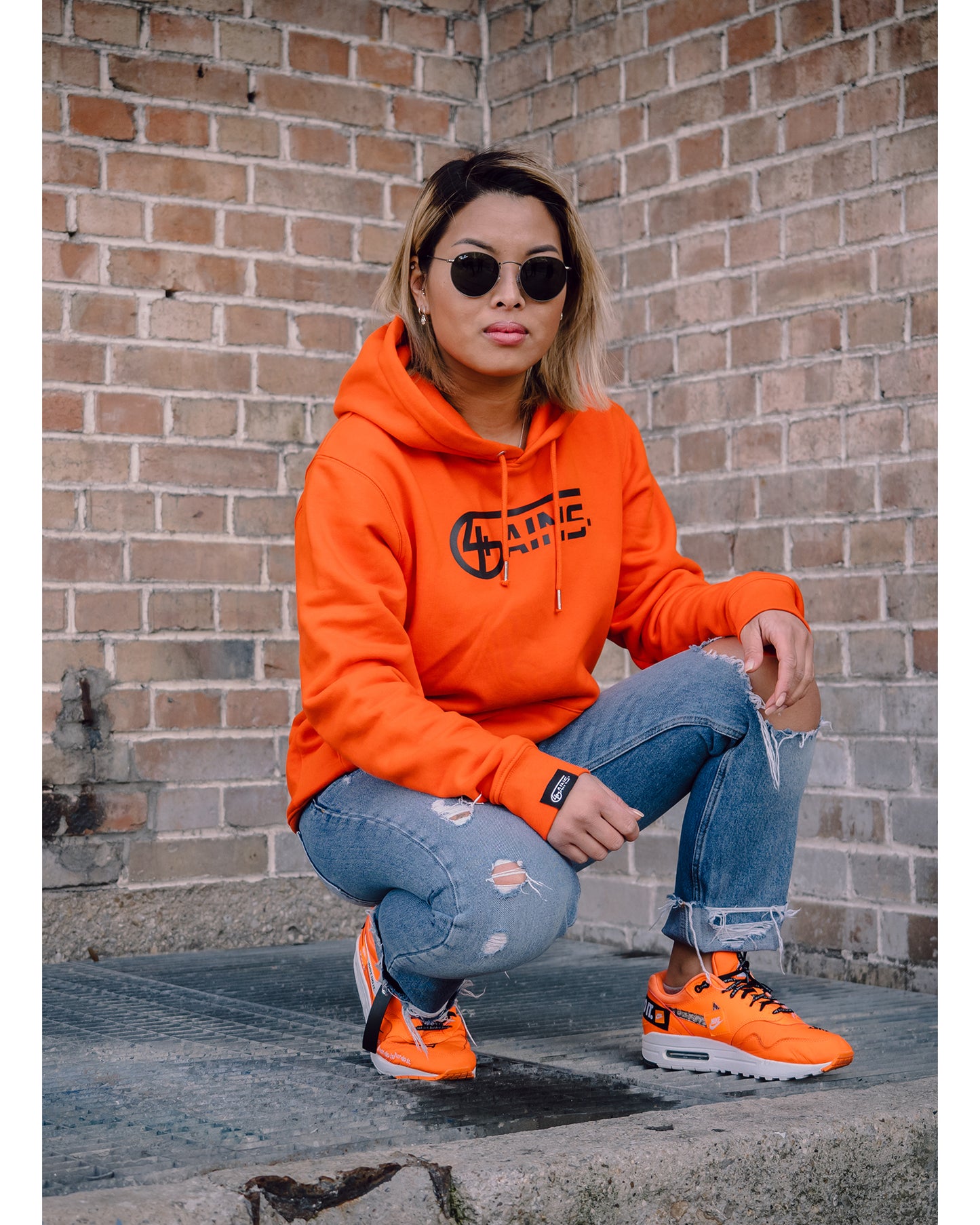 Weibliches Model kniet hin mit 4GAINS Hoodie in orange/black