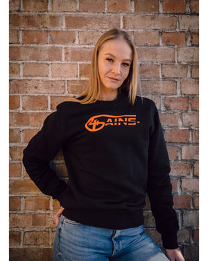 Weibliches Model steht an der Wand und hat 4GAINS unisex Sweater an in black/orange