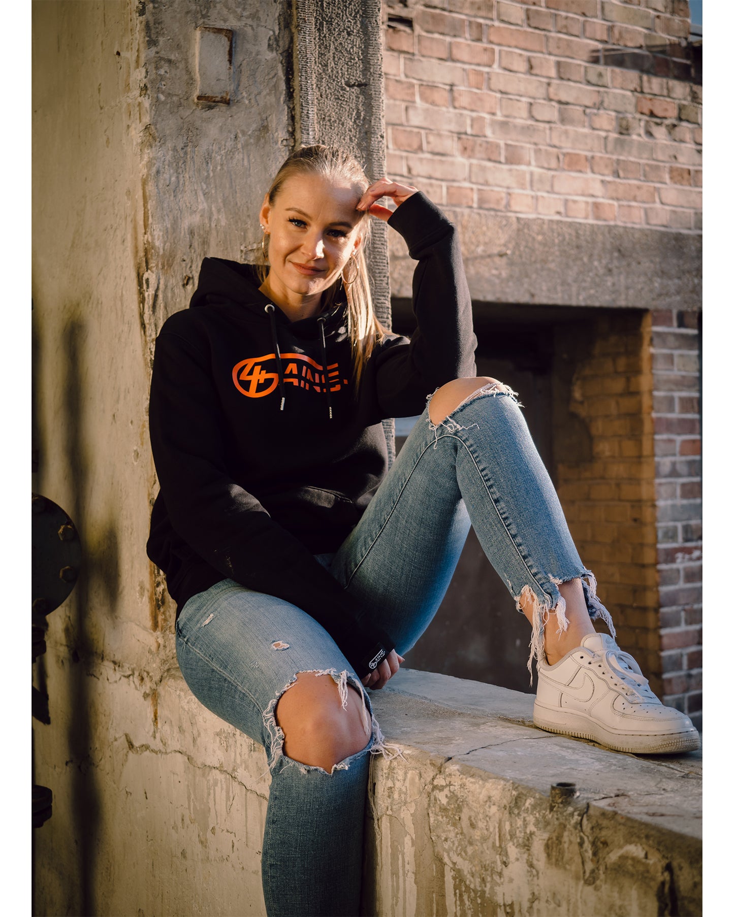 Weibliches Model sitzt an der Wand und hat 4GAINS unisex Hoodie an in black/orange