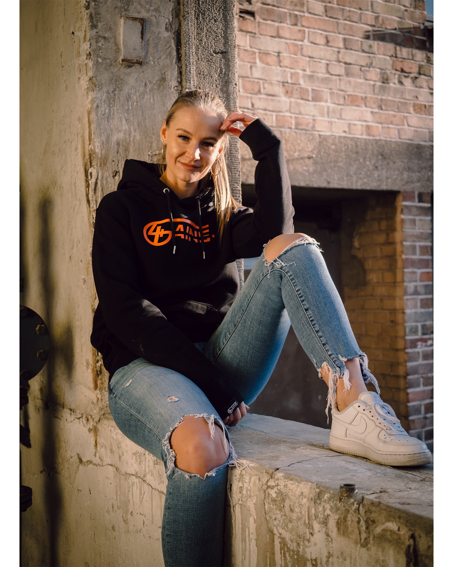 Weibliches Model sitzt an der Wand und hat 4GAINS unisex Hoodie an in black/orange