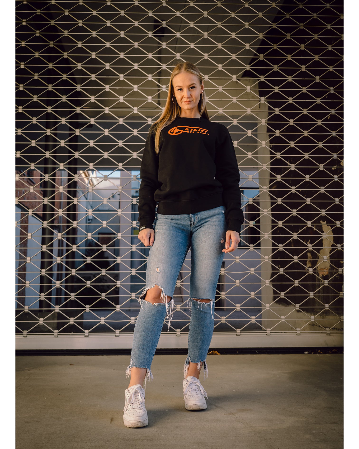 Weibliches Model steht vor einem Rolltor und hat 4GAINS unisex Sweater an in black/orange