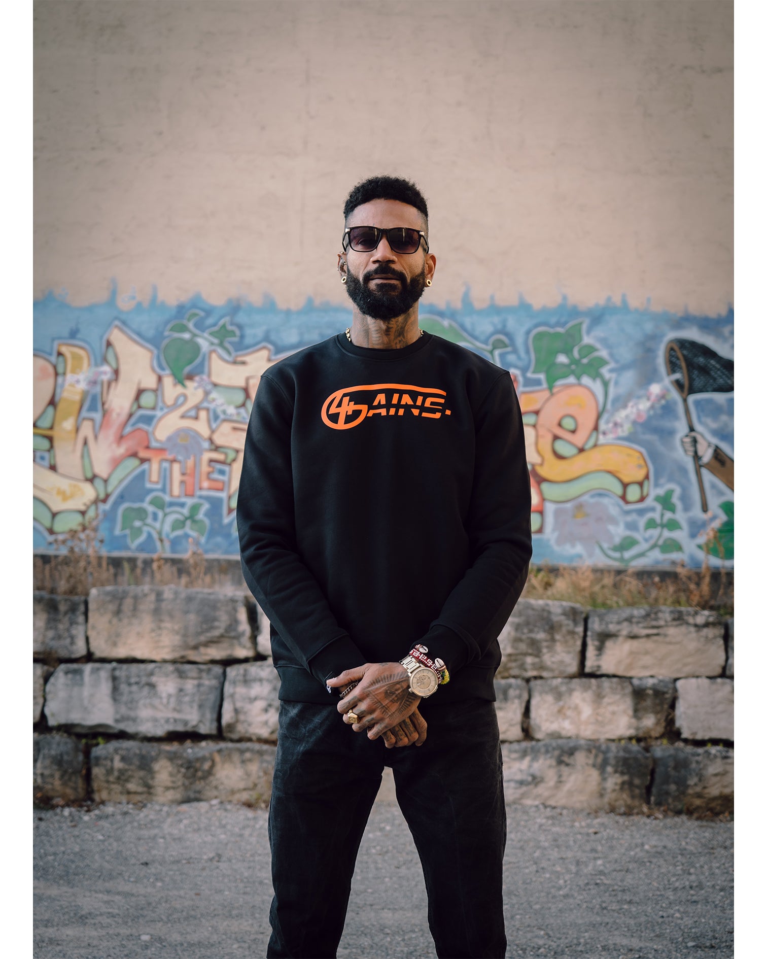 Männliches Model steht vor Graffitiwand und hat 4GAINS unisex Sweater an in black/orange