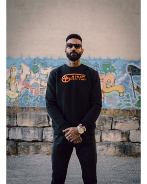Männliches Model steht vor Graffitiwand und hat 4GAINS unisex Sweater an in black/orange