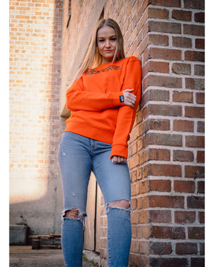Weibliches Model steht an der Wand und hat 4GAINS unisex Sweater an in orange/black