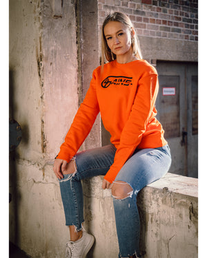 Weibliches Model sitzt an der Wand und hat 4GAINS unisex Sweater an in orange/black