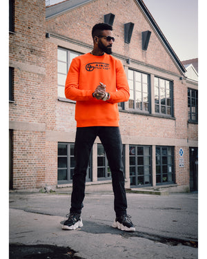 Männliches Model steht  und hat 4GAINS unisex Sweater an in orange/black