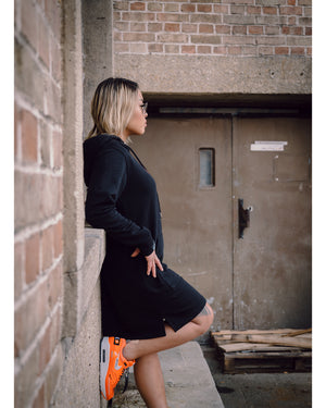 Weibliches Model steht vor einer Wand mit orangenen Schuhen und dem 4GAINS Sweaterkleid für Frauen in black/orange