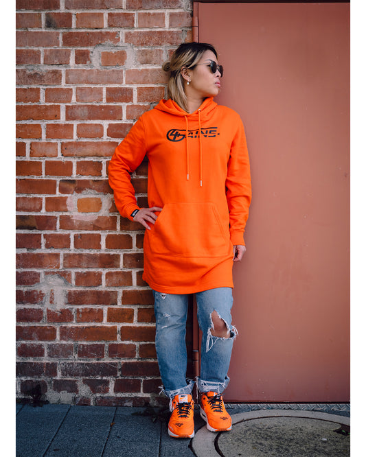 Weibliches Model steht vor einer Wand mit orangenen Schuhen und dem 4GAINS Sweaterkleid für Frauen in orange/black