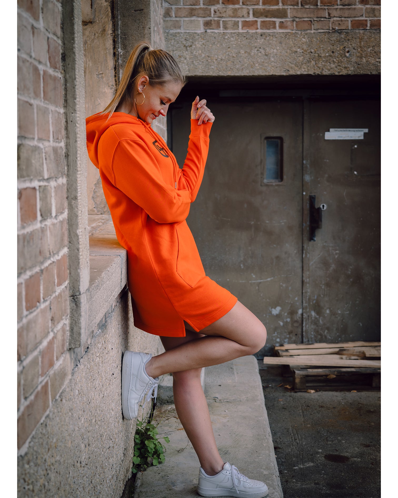 Weibliches Model steht vor einer Wand mit weißen Schuhen und dem 4GAINS Sweaterkleid für Frauen in orange/black